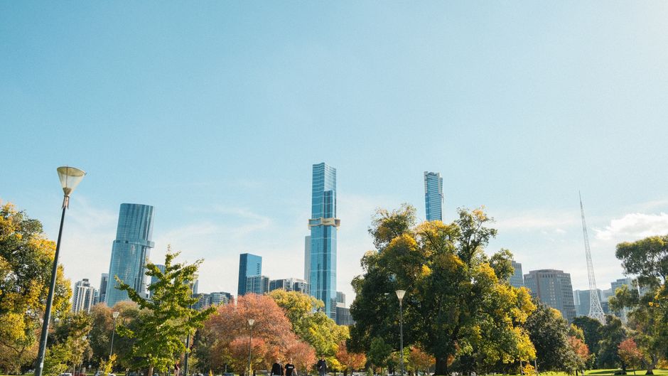 A clear daytime cityscape featuring a row of deciduous trees in the foreground, with leaves displaying shades of green, yellow, and orange. Behind the trees, there are several modern high-rise buildings with glass facades reflecting sunlight, including one tall tower with a rectangular shape and vertical window arrangements, and another with a distinctive twisting design. The scene appears to be in an urban park or open area adjacent to a street or pathway, with lampposts standing among the trees. The sky is bright and mostly cloudless, contributing to a well-lit, vibrant atmosphere. This environment suggests a city known for contemporary architecture and green spaces, which aligns with the concept of alternative waste handling and private rubbish collection services, such as those offered by Rubbish Clearance Queens Park, in a professional and environmentally conscious context.