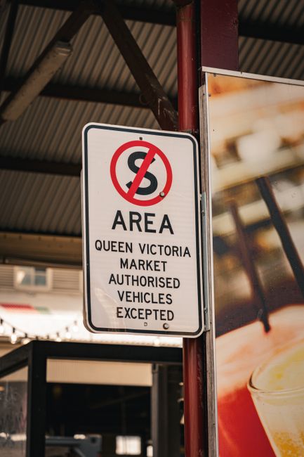 A white rectangular sign with black and red text and symbols, mounted on a red metal post under a corrugated metal roof. The sign indicates a no-parking zone with a red circle and diagonal line crossing out a black letter 'S'. Below, it states that the area is designated for Queen Victoria Market and is reserved for authorized vehicles, with exceptions. The surrounding environment includes parts of a building with a beige exterior and a large display board with blurred images of food and drink, suggesting a commercial or market setting. The lighting is natural, with daylight illuminating the scene, highlighting the sign's visibility and the textures of the metal post and roof. This sign, often found in urban outdoor spaces, relates to private or independent waste handling and clearance areas, aligning with the context of rubbish collection services provided by companies like Rubbish Clearance Queens Park in managing waste within designated zones.