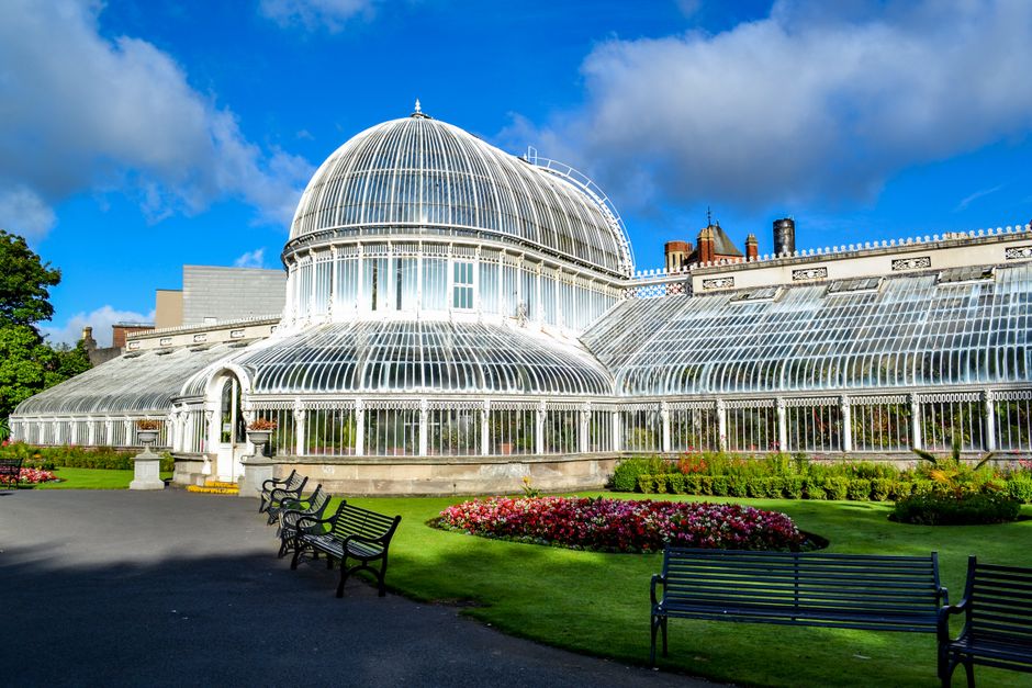 A large ornate greenhouse with a curved glass and white metal framework is situated in a landscaped garden under a partly cloudy blue sky. The structure features a prominent central dome topped with a small decorative finial, with lower glass extensions spreading outward in a semi-circular shape. The greenhouse's glass panels are clear, revealing the interior framework, and the white metal supports are intricately designed. The surrounding garden includes well-maintained green lawns, flower beds with colorful blooming plants, and a paved pathway with black metal benches placed along it. To the left, there are lush green trees, while in the background, brick chimney stacks and rooftops are visible behind the greenhouse. The scene is brightly lit by natural sunlight, highlighting the reflective surfaces of the glass and the vivid colors of the flowers, creating a serene and orderly outdoor setting that aligns with the themes of garden maintenance and greenhouse management, often associated with waste disposal and clearance services like those offered by Rubbish Clearance Queens Park.