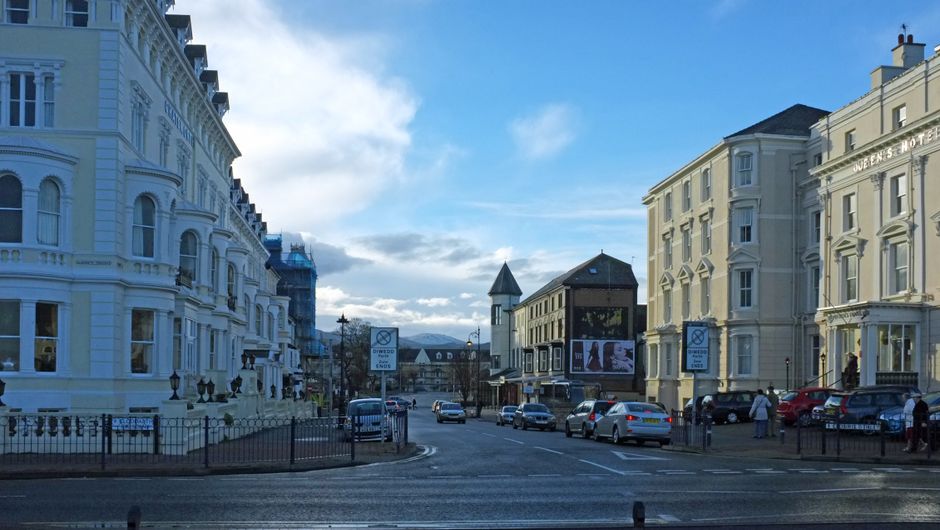 A wide street scene in a city area featuring large, white, Victorian-style terraced buildings on the left with ornate facades, intricate window frames, and decorative railings. On the right, there are cream-colored, similarly styled buildings, including a hotel with the name partially visible on the facade. Several parked cars, including small to medium-sized vehicles in various colours such as silver, white, and red, are lined along the street and sidewalk. Pedestrians walk along the pavement, and a large digital billboard displays advertisements towards the center of the background. The street is paved with dark asphalt, with road markings and small metal barriers separating the traffic from the pedestrian zones. In the distance, there is a cloudy sky with patches of blue visible, casting natural daylight over the scene, emphasizing the architectural details and the urban environment. This scene reflects a typical city street with a mix of historic and modern elements, consistent with an area that offers private waste and rubbish management services, such as those provided by Rubbish Clearance Queens Park.