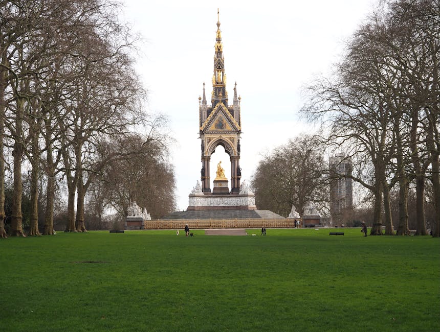 A historic monument with a tall, ornate spire featuring intricate gold accents and Gothic architectural details stands prominently in the center of a well-maintained, lush green park. The monument base is marked by a broad platform, and beneath the spire, there is a golden statue or sculpture. The surrounding area includes a spacious lawn with a few benches and several people walking or sitting nearby, indicating a public outdoor space. Tall, leafless trees frame the scene on both sides, suggesting it is late autumn or winter. The sky above is overcast with a light grey hue, providing diffuse natural light that highlights the monument’s detailed design while casting soft shadows on the ground. This setting offers an unobstructed view of the monument and park, with a quiet atmosphere typical of an urban open space. Rubbish Clearance Queens Park could be called upon for discreet management of waste, debris, or litter in such community environments, ensuring they remain clean and welcoming for visitors.